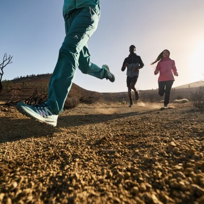 Drei Menschen laufen auf einem unbefestigten Weg in einer trockenen, hügeligen Landschaft.