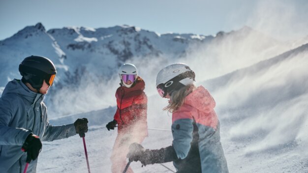 Drei Skifahrende in Helmen und Winterjacken stehen auf einer verschneiten Bergpiste, Pulverschnee wirbelt auf, sonnige Berggipfel im Hintergrund. | © INTERSPORT International Corporation GmbH