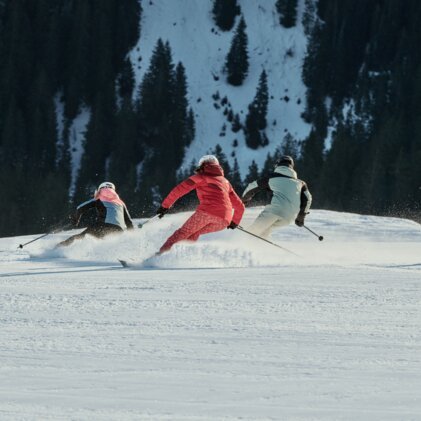 Drei Skifahrende in bunten Jacken schneiden parallele Schwünge auf einer präparierten Piste, Tannenwald und schneebedeckte Bergflanke im Hintergrund. | © INTERSPORT International Corporation GmbH