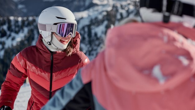 Lächelnde Skifahrerin mit weißem Helm und rosa Skibrille in roter Jacke; im Vordergrund unscharfe Begleitperson und verschneite Berglandschaft im Hintergrund. | © INTERSPORT International Corporation GmbH
