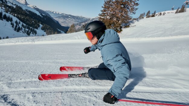 Skifahrer in blauem Anorak und schwarzem Helm mit roten Skiern macht eine Carving-Kurve auf präparierter Piste vor verschneiten Bergen. | © INTERSPORT International Corporation GmbH