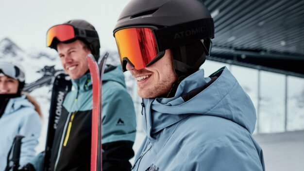 Drei Skifahrer in Winterjacken und Helmen; im Vordergrund ein lächelnder Mann mit schwarzem Helm und roten Skibrillen, hält rote Ski, verschneite Bergkulisse im Hintergrund. | © INTERSPORT International Corporation GmbH
