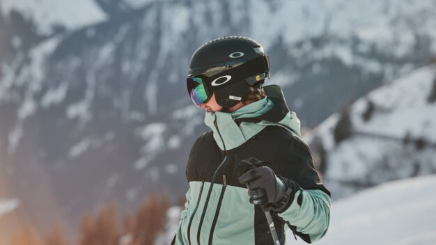 Skifahrer in schwarzem Helm mit verspiegelter Oakley-Skibrille und mint-schwarzer Jacke, mit Handschuhen einen Skistock haltend vor verschneiter Berglandschaft. | © INTERSPORT International Corporation GmbH