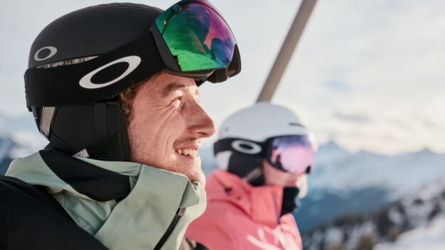 Zwei Skifahrer auf einem Sessellift: Nahaufnahme eines lächelnden Mannes mit schwarzem Helm und verspiegelten Skibrillen, im Hintergrund eine Frau in weißem Helm und rosa Skijacke vor schneebedeckten Bergen. | © INTERSPORT International Corporation GmbH