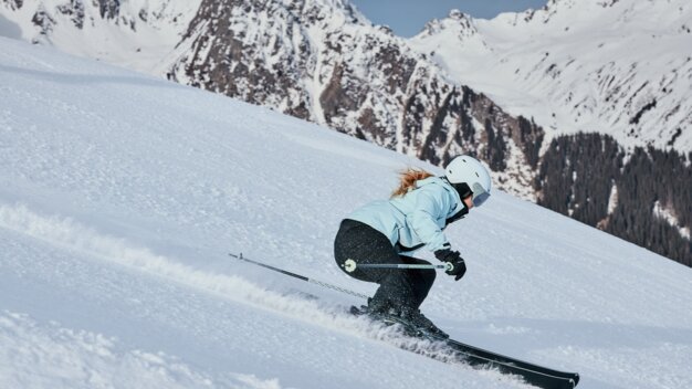 Frau beim Skifahren in hellblauer Jacke und weißem Helm, sie fährt eine steile, verschneite Piste vor schneebedeckten Bergen. | © INTERSPORT International Corporation GmbH