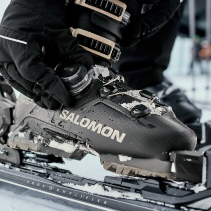 Genaue Nahaufnahme einer Hand mit Ski­handschuh, die eine verschneite Salomon-Skischuh­schnalle auf einer gebundenen Ski­bindung einstellt. | © INTERSPORT International Corporation GmbH