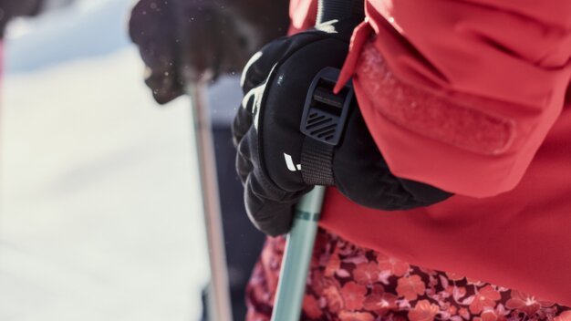 Nahaufnahme einer schwarz behandschuhten Hand, die einen Skistock hält; rote Skijacke und geblümte Skihose, verschneite Berglandschaft im Hintergrund. | © INTERSPORT International Corporation GmbH