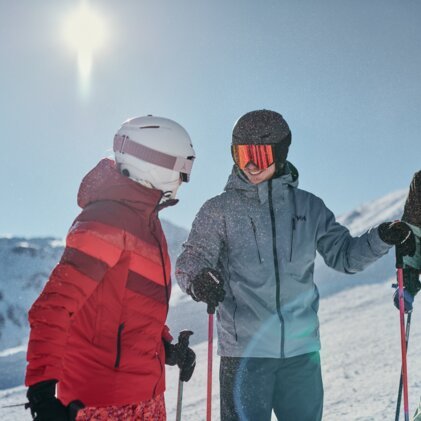 Drei Skifahrer in Winterjacken und Helmen stehen auf verschneiter Piste, lachen und stützen sich auf Skistöcke; sonnige Bergkulisse im Hintergrund. | © INTERSPORT International Corporation GmbH