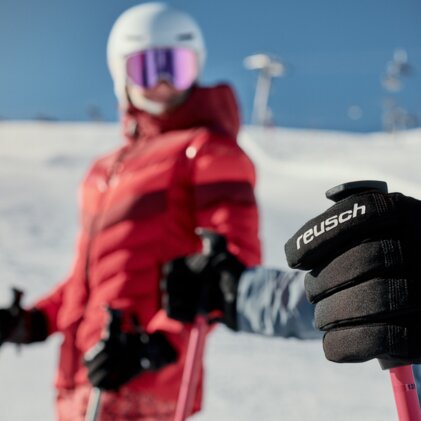 Nahaufnahme einer Hand mit schwarzem Reusch-Skihandschuh, die einen Skistock hält; im unscharfen Hintergrund eine Person in roter Skijacke und Helm auf verschneiter Piste. | © INTERSPORT International Corporation GmbH
