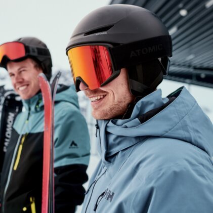 Drei Skifahrer in Winterjacken und Helmen; im Vordergrund ein lächelnder Mann mit schwarzem Helm und roten Skibrillen, hält rote Ski, verschneite Bergkulisse im Hintergrund. | © INTERSPORT International Corporation GmbH