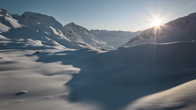 Verschneites alpines Tal bei Sonnenaufgang mit sonnenbeschienenen Berggipfeln, sanften Schneehügeln und einer Spur von Fußabdrücken im Vordergrund. | © INTERSPORT International Corporation GmbH