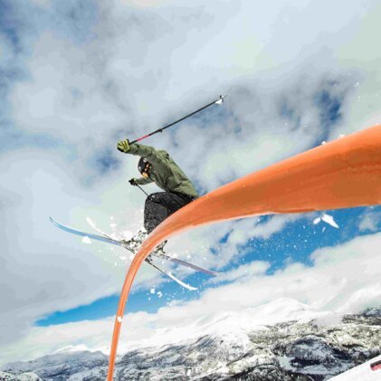 Skifahrer in grüner Jacke und Helm springt über ein gebogenes orangefarbenes Rohr im Snowpark; Schnee, Berge und bewölkter Himmel im Hintergrund. | © INTERSPORT International Corporation GmbH
