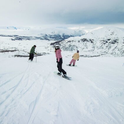 Drei Personen beim Snowboarden auf einer weiten, verschneiten Piste mit schneebedeckten Bergen im Hintergrund und bewölktem Himmel. | © INTERSPORT International Corporation GmbH