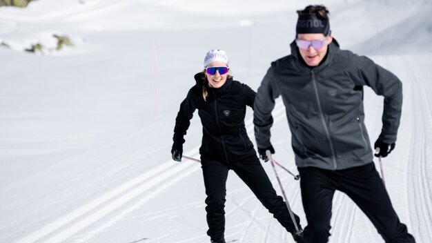 Zwei Langläufer in schwarzer Outdoor-Bekleidung und Schutzbrillen auf einer präparierten Loipe im Sonnenschein, die hintereinander skiend unterwegs sind, Frau lächelt. | © INTERSPORT International Corporation GmbH