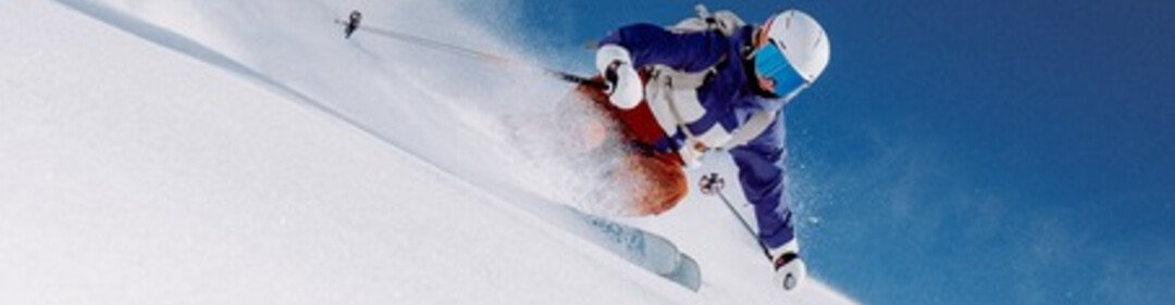 Skifahrer in blauem Jacket und weißem Helm fährt diagonal eine steile Pulverschneepiste, Schneestaub spritzt. | © INTERSPORT International Corporation GmbH