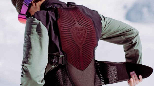 Person von hinten, die einen gepolsterten Rückenprotektor/Airbag‑Weste über orangefarbene Skihose anlegt; trägt grüne Jacke, braune Mütze und lila Skibrille vor verschneiter Berglandschaft. | © INTERSPORT International Corporation GmbH