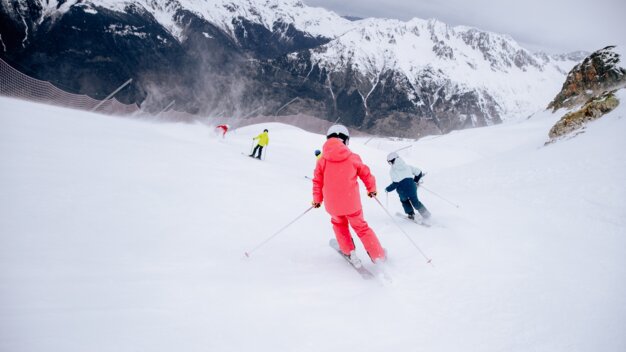 Drei Skifahrer auf einem verschneiten Hang, vorn eine Person in roter Skijacke und Helm mit Skistöcken, im Hintergrund schneebedeckte Berge und Sicherheitsnetz. | © INTERSPORT International Corporation GmbH