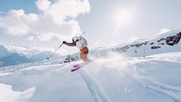 Skifahrer in weißer Jacke und orangener Hose springt mit pinken Skiern im Tiefschnee auf sonnigem Alpenhang. | © INTERSPORT International Corporation GmbH