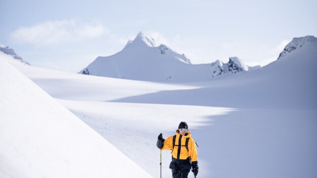 Skitourengeher in gelber Jacke mit Skistöcken in weiter, verschneiter Berglandschaft vor schneebedeckten Gipfeln. | © INTERSPORT International Corporation GmbH