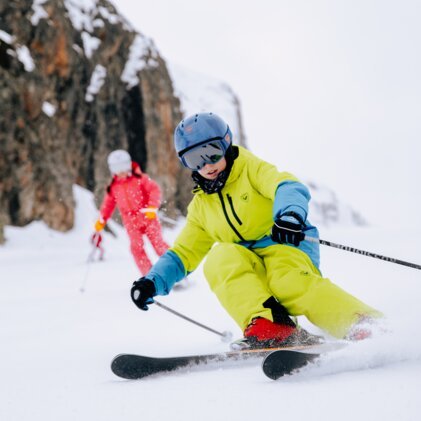 Kind beim Skifahren: neongelber Skianzug, blauer Helm und rote Skischuhe schneidet Schwung auf Piste, zweite Person in rotem Anzug im Hintergrund vor Felswand. | © INTERSPORT International Corporation GmbH