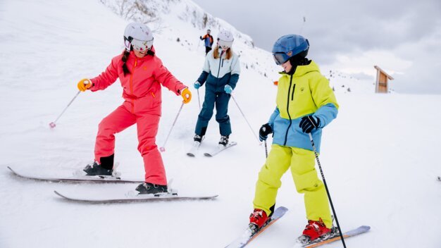 Drei Kinder in bunten Skianzügen (rot, gelb, blau) mit Helmen und Skistöcken beim Skifahren auf einer verschneiten Piste vor bergigem Hintergrund. | © INTERSPORT International Corporation GmbH