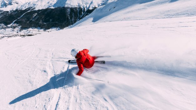 Skifahrer in roter Jacke und weißem Helm beim Carven den Hang hinab, Pulverschnee sprüht, verschneite Bergkulisse im Hintergrund. | © INTERSPORT International Corporation GmbH