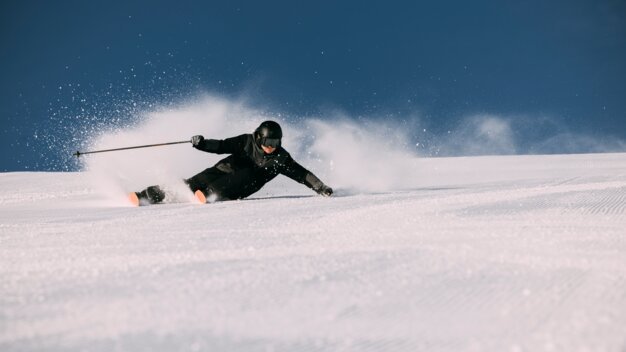 Skifahrer in schwarzer Ausrüstung und Helm mit orangefarbenen Skispitzen beim Carving, Schneespray auf präparierter Piste vor klarem blauem Himmel. | © INTERSPORT International Corporation GmbH