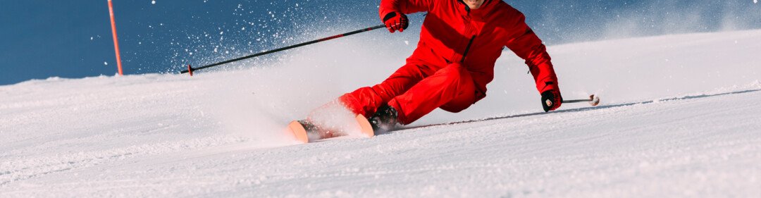 Skifahrer in roter Jacke und Helm fährt dynamisch eine präparierte Piste hinunter, Schnee spritzt, blauer Himmel im Hintergrund. | © INTERSPORT International Corporation GmbH