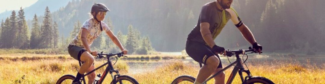 Zwei Radfahrer auf einer Wiese im warmen Abendlicht, umgeben von Bergen und Wald in einer natürlichen Landschaft.