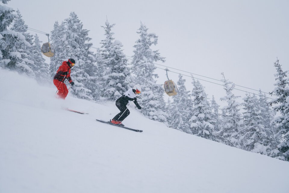 © INTERSPORT International Corporation GmbH Zwei Skifahrer fahren auf einer schneebedeckten Piste unter einer Gondelbahn hindurch. | © INTERSPORT International Corporation GmbH