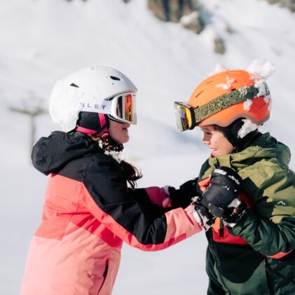 Zwei Kinder in Skikleidung und Helmen, die sich auf der Piste ansehen.