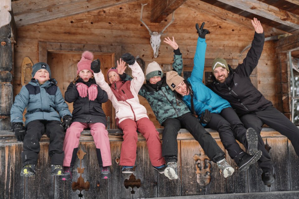 © INTERSPORT International Corporation GmbH Gruppe von Personen in Winterkleidung sitzt vor einer Holzhütte und winkt. | © INTERSPORT International Corporation GmbH
