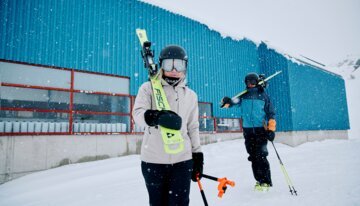 © Fischer Sports GmbH Zwei Skifahrer mit Skiern vor blauem Gebäude im Schnee. | © Fischer Sports GmbH