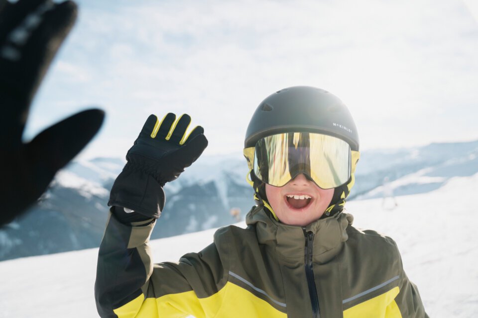 © INTERSPORT International Corporation GmbH Junge mit Skibrille und Helm winkt auf einer Skipiste. | © INTERSPORT International Corporation GmbH