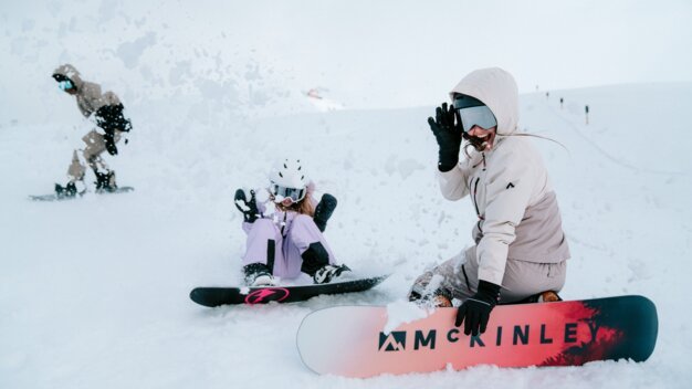 Zwei Snowboarder und eine Person im Hintergrund auf einem verschneiten Hang.